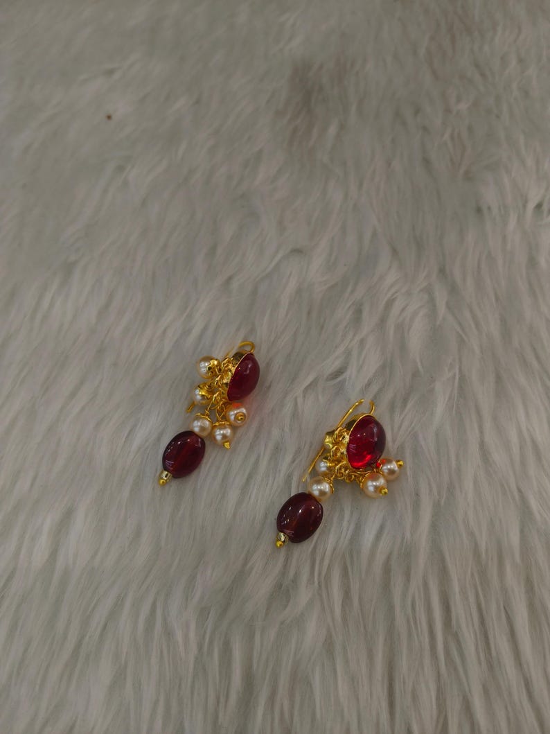 May include: A pair of gold-toned earrings featuring a large, oval-shaped deep red stone at the top, accented by smaller white pearls and a dangling dark red oval bead at the bottom, suspended from a gold-toned hook mechanism for ear attachment.