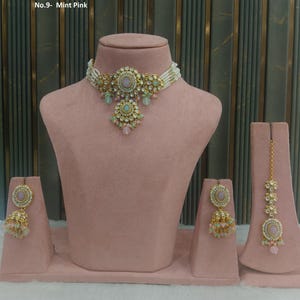 May include: A mint pink jewelry set displayed on a pink mannequin. The set includes a necklace, earrings, and a headpiece. The necklace and earrings have a gold-colored metal with pink and green accents. The headpiece has a gold chain.