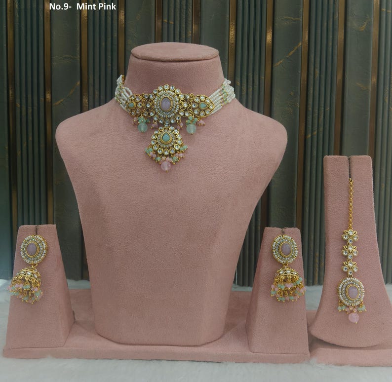 May include: A mint pink jewelry set displayed on a pink mannequin. The set includes a necklace, earrings, and a headpiece. The necklace and earrings have a gold-colored metal with pink and green accents. The headpiece has a gold chain.