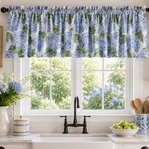 May include: A window valance featuring a repeating pattern of blue hydrangea blossoms and green leaves on a white background. The fabric is gathered along a dark rod, creating a ruffled top edge that hangs above a kitchen window frame.