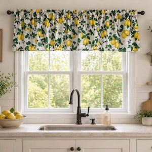May include: A white window valance with a lemon and leaf pattern. The fabric features yellow lemons, green leaves, and small blue berries. The valance is mounted above a double window in a kitchen setting.