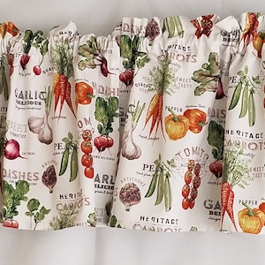 Op de afbeelding: Een witte stoffen valance met een herhalend patroon van verschillende groenten, waaronder wortelen, tomaten, knoflook en artisjokken. De valance heeft de tekst "Heritage Carrots" en "Garlic Delicious".