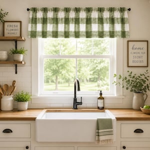 May include: A green and white buffalo check window valance hangs above a white farmhouse sink with a black faucet. The kitchen features wooden countertops, white cabinets, and various decorative items including signs that read Fresh Flowers Market.