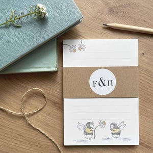 May include: A stack of white notepads with a brown paper band featuring the initials "F & H" in a white circle. The notepads have a bee and daisy illustration. A light blue book and pencil are in the background.