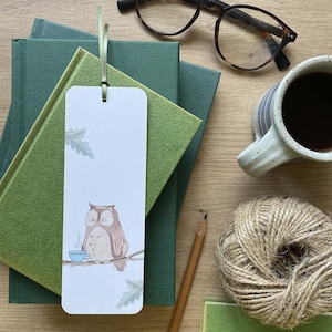 May include: A white bookmark featuring an illustration of a brown owl holding a blue teacup. The owl is perched on a branch with green leaves. The bookmark is placed on top of a stack of green books.