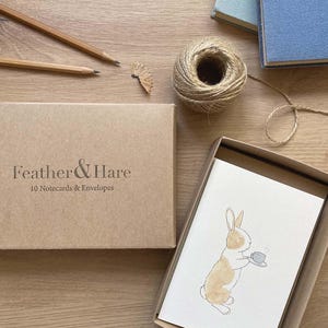 May include: A brown cardboard box labelled Feather & Hare 10 Notecards & Envelopes next to an open box containing a white notecard with a watercolour illustration of a light brown rabbit holding a small grey teacup and saucer.