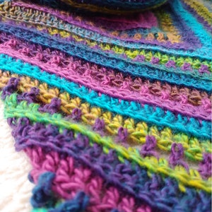 Pode incluir: Uma echarpe de croch&ecirc; colorida com um padr&atilde;o listrado em tons de roxo, azul, verde e amarelo. A echarpe tem franjas na ponta.