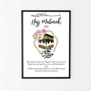 May include: A framed print with the words "Hajj Mubarak 2023" in elegant script, featuring an illustration of the Kaaba, floral accents, and Arabic calligraphy. The print is in a black frame.