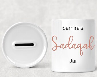 Puede incluir: Hucha de cerámica blanca con una ranura en la parte superior. La hucha tiene el texto "Samira's Sadaqah Jar" escrito en ella en una fuente cursiva rosa.