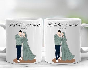 Op de afbeelding: Twee witte keramische mokken met zwarte handvatten. Elke mok heeft een cartoonillustratie van een stel dat elkaars hand vasthoudt. De tekst "Habibi Ahmed 12/02/23" staat op de ene mok en "Habibi Zainab 12/02/23" op de andere mok.