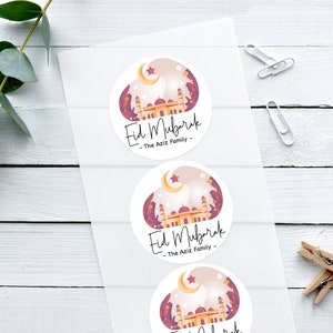 Può includere: Tre adesivi rotondi con sfondo bianco e un'illustrazione colorata di una moschea con una mezzaluna e una stella. Il testo "Eid Mubarak - The Aziz Family -" è stampato su ogni adesivo.