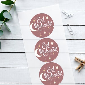 Può includere: Tre adesivi rotondi con sfondo rosa e testo bianco che dice "Eid Mubarak". Gli adesivi hanno una mezzaluna e stelle dorate.
