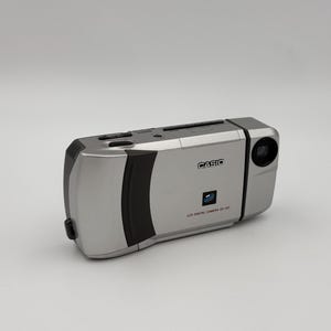 May include: A silver Casio LCD digital camera with a built-in lens. The camera has a black rubber grip and a small LCD screen on the back.