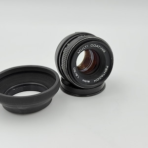 May include: A black Pentacon 1.8/50 auto camera lens with a matching black lens hood. The lens has the words "Multi Coating" and "Pentacon" printed on it. The lens hood is round and has a ridged design.