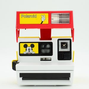 Könnte beinhalten: Vintage Polaroid-Kamera mit rotem und gelbem Design. Die Kamera hat das Wort "Polaroid" in Gelb, eine Mickey Mouse-Grafik und einen Blitz. Die Kamera ist weiß mit schwarzen Akzenten.