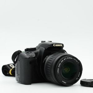 Op de afbeelding: Een zwarte Canon DSLR-camera met een lens bevestigd en een lensdop. De camerariem is zwart met gele en witte tekst. De lens heeft de tekst "Canon Zoom Lens EF-S 18-55mm 1:3.5-5.6 II".