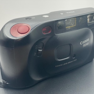 Op de afbeelding: Een zwarte Canon Prima camera met een rode knop en een lens met de tekst "Canon Lens 35mm 1:4.5".