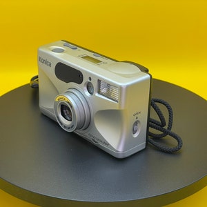 以下が含まれることがあります： 黒いストラップが付いた銀色のコニカ・センチュリア300カメラ。カメラには「コニカレンズ35-80mm」と書かれたレンズカバーが付いています。