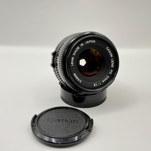 May include: A black Canon FD 50mm 1:1.8 lens with a black lens cap. The lens is made in Japan and has the words "Canon Lens FD 50mm 1:1.8" printed on it.