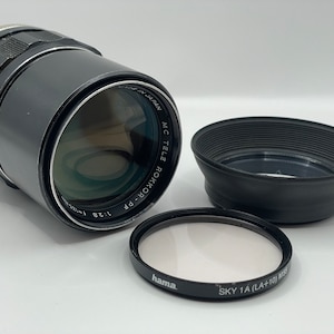 May include: A silver and black camera lens with the text "MC TELE ROKKOR-PP 1:28" printed on the side. The lens is accompanied by a black lens hood and a filter.