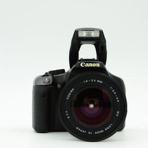 May include: Black Canon camera with a Cosina 19-35 mm lens. The lens has a red interior and the camera has a built-in flash. The camera is set against a white background.