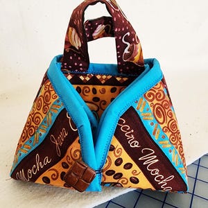 May include: A quilted coffee cup cozy with a handle. The cozy features a brown, yellow, and blue patterned fabric with the words "Mocha Java" and "Cappuccino Mocha". A brown square button secures the cozy.
