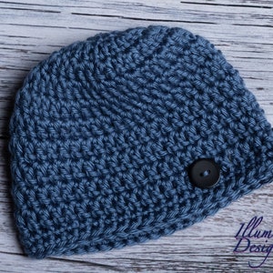 May include: A blue crocheted beanie with a black button on the side. The beanie is on a white wooden background.