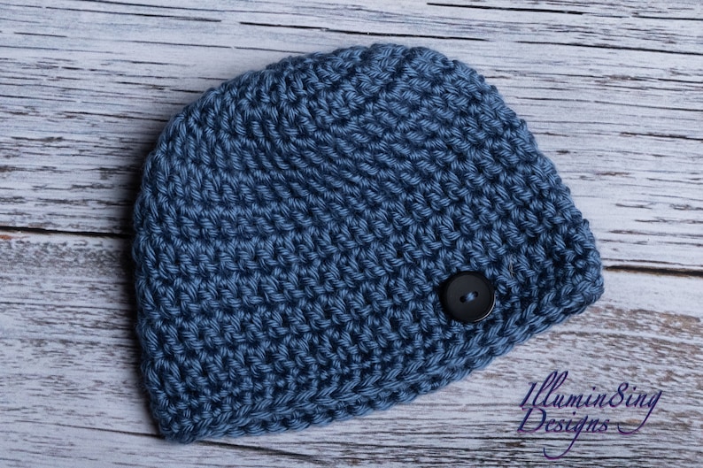May include: A blue crocheted beanie with a black button on the side. The beanie is on a white wooden background.
