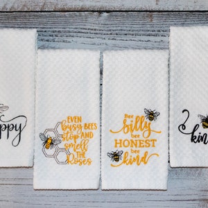 May include: Set of four white kitchen towels with embroidered bees and bee-themed sayings. The towels feature the phrases "happy", "Even busy bees stop and smell the roses", "Bee silly bee honest bee kind", and "Bee kind".