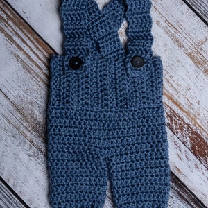 May include: Blue crocheted baby overalls with black buttons. The overalls are on a white wood background.