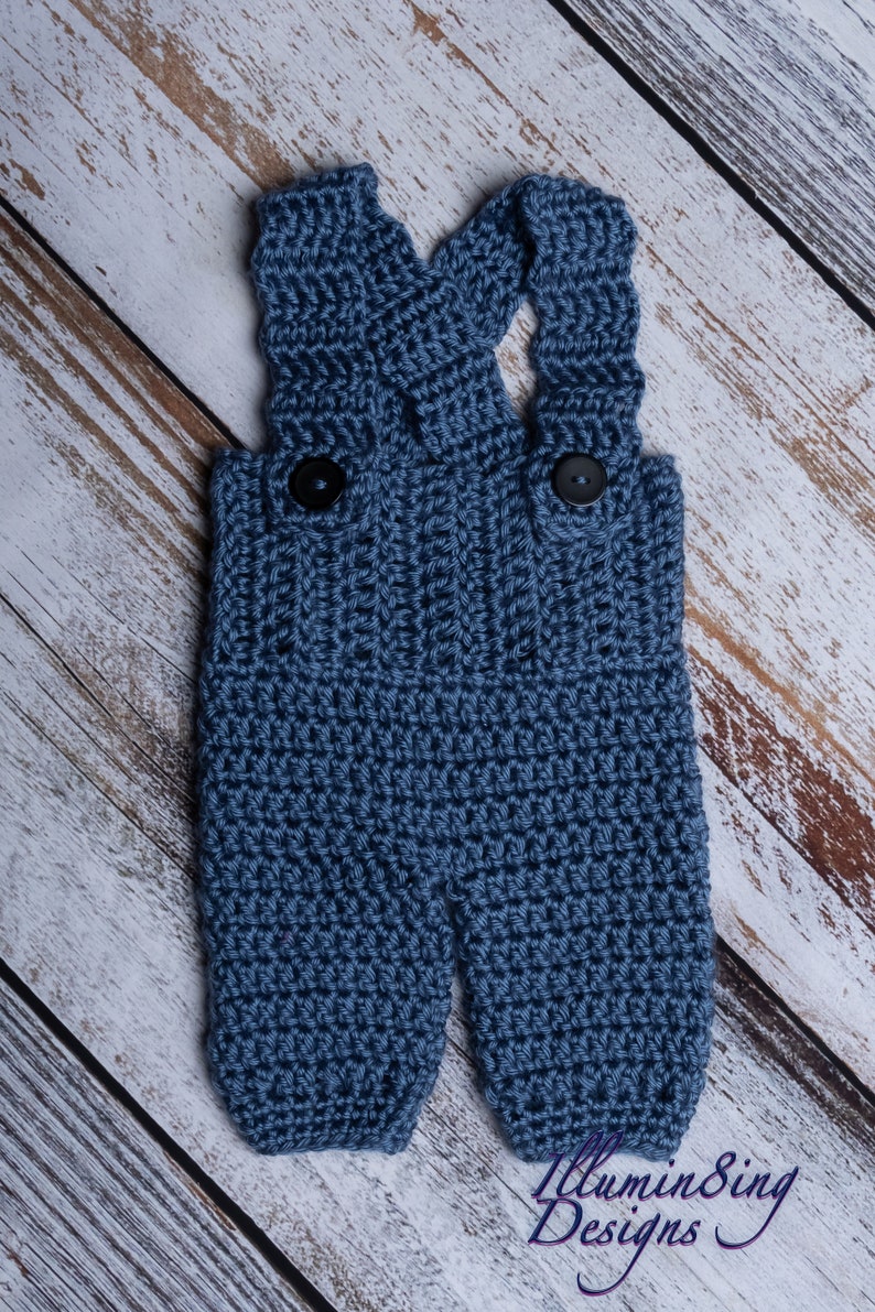 May include: Blue crocheted baby overalls with black buttons. The overalls are on a white wood background.