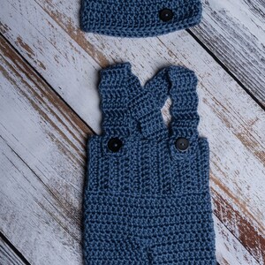 May include: A blue crocheted baby outfit with a matching hat. The outfit features a pair of overalls with straps and two buttons. The hat is a simple beanie style with a button on top.