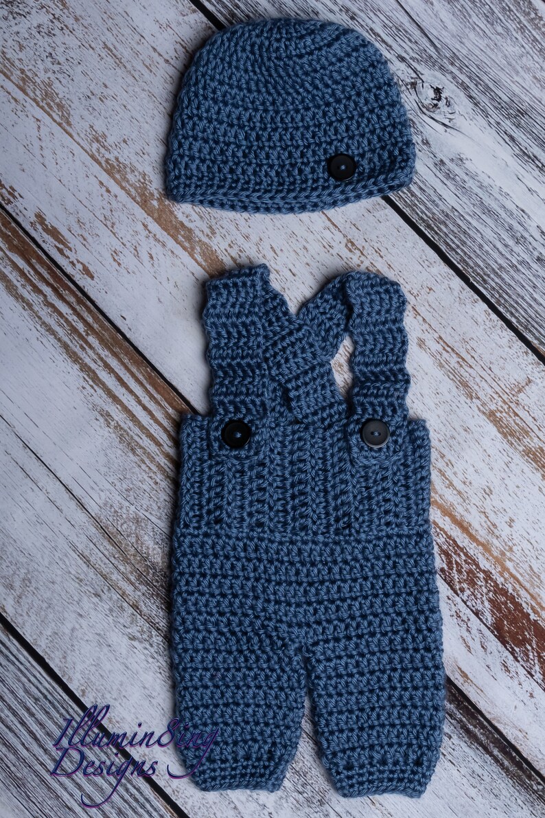 May include: A blue crocheted baby outfit with a matching hat. The outfit features a pair of overalls with straps and two buttons. The hat is a simple beanie style with a button on top.