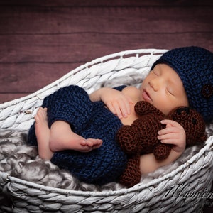 May include: A newborn baby wearing a blue knitted hat and matching pants sleeps in a white wicker basket. The baby is holding a brown knitted teddy bear.
