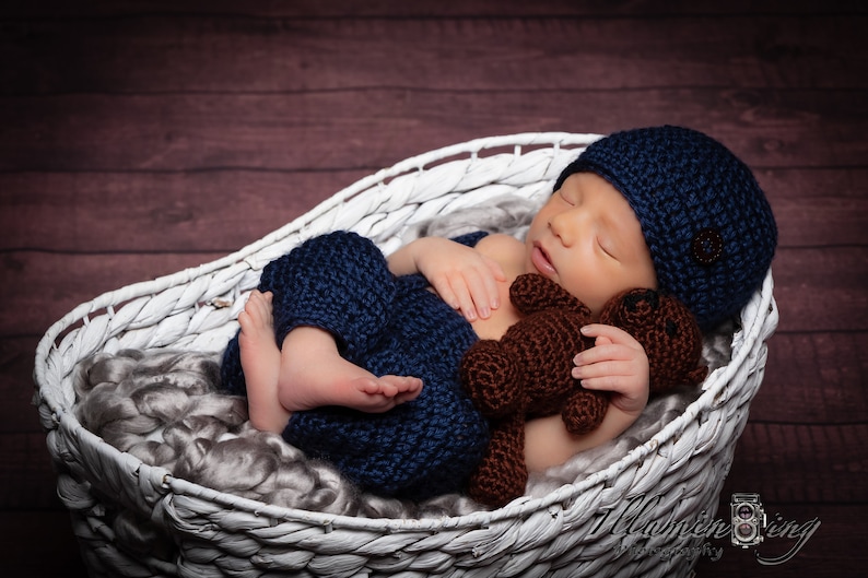 May include: A newborn baby wearing a blue knitted hat and matching pants sleeps in a white wicker basket. The baby is holding a brown knitted teddy bear.