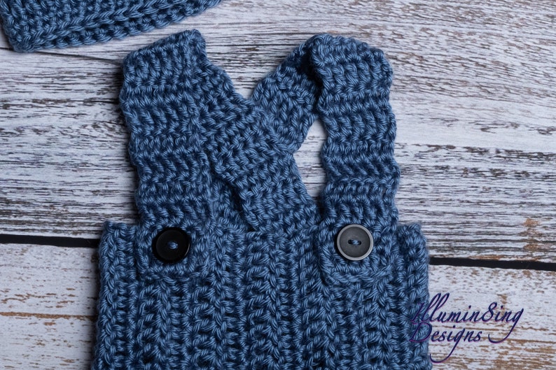 May include: Blue knitted baby overalls with black buttons. The overalls have a criss-cross strap design in the back.