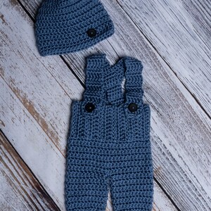 May include: A blue crocheted baby outfit with a matching hat. The outfit features a pair of overalls with two black buttons and the hat has a single black button.
