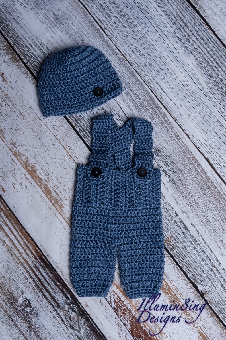 May include: A blue crocheted baby outfit with a matching hat. The outfit features a pair of overalls with two black buttons and the hat has a single black button.