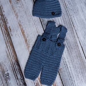 May include: A blue crocheted baby outfit with a matching hat. The outfit features a pair of overalls with a button closure and the hat has a simple design.