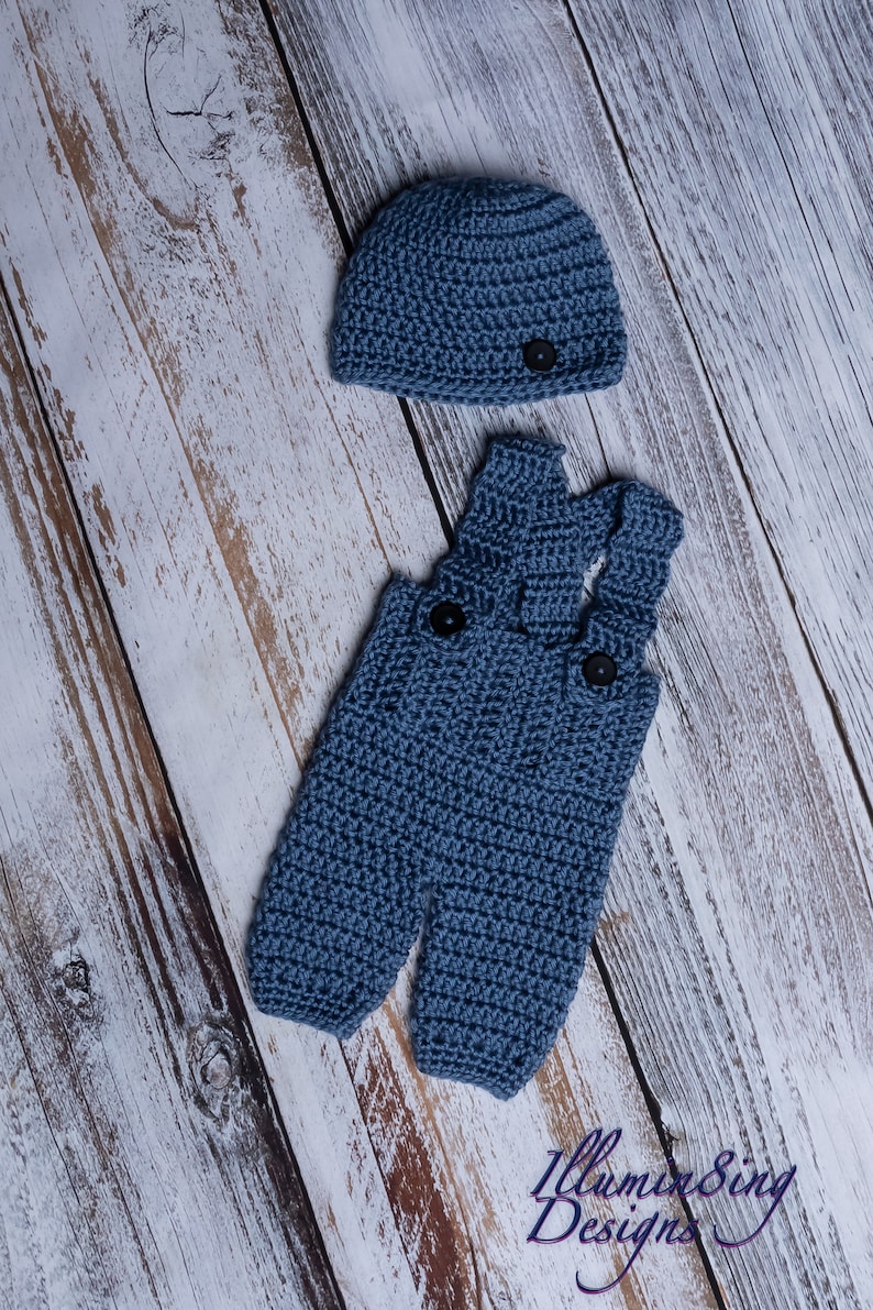 May include: A blue crocheted baby outfit with a matching hat. The outfit features a pair of overalls with a button closure and the hat has a simple design.