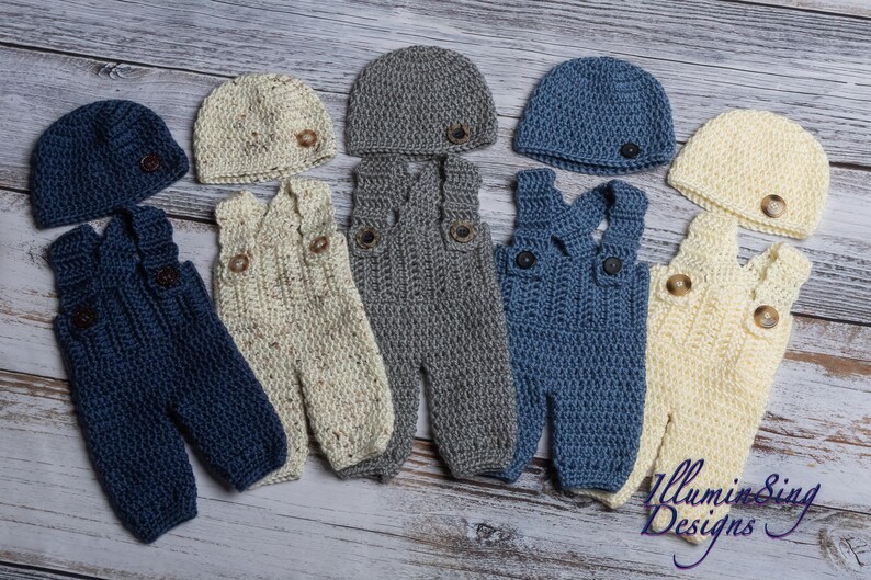 May include: Five sets of crocheted baby overalls and hats in different colors. The overalls are in blue, cream, and gray. The hats are in the same colors. Each set has a button on the hat and two buttons on the overalls.