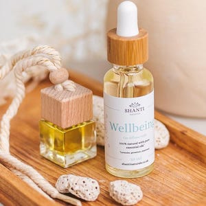 May include: A glass bottle with a wooden cap and a label that reads "Wellbeing Car diffuser refill 100% natural with pure essential oils Lavender, geranium, marjoram 30 ml shantinaturals.co.uk". The bottle is sitting on a wooden tray with a white rope and two white stones.