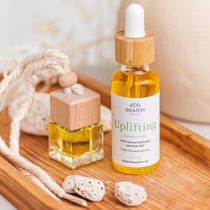 Puede incluir: Un frasco difusor de coche de vidrio transparente con tapa de madera y cuerda, junto a un frasco cuentagotas de vidrio ámbar de 30 ml con la etiqueta Shanti Naturals Uplifting, que contiene aceites esenciales de naranja, limoncillo y pomelo.