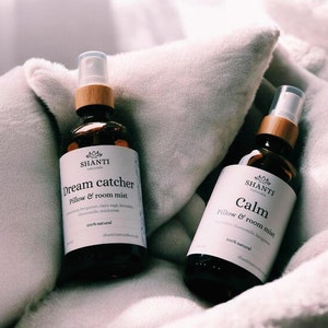 Könnte beinhalten: Zwei braune Glassprühflaschen mit Holzaufsätzen, beschriftet mit "Dream Catcher" und "Calm" Kissen- und Raumspray. Die Flaschen stehen auf einer weichen, weißen, strukturierten Oberfläche. Die Etiketten tragen den Markennamen "SHANTI naturale".