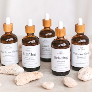 May include: Five amber glass massage oil bottles with white labels and wooden caps. The labels read "Muscle Soothing", "Uplifting", "Love", "Relaxing", and "Womens blend". The bottles are surrounded by light-colored stones.
