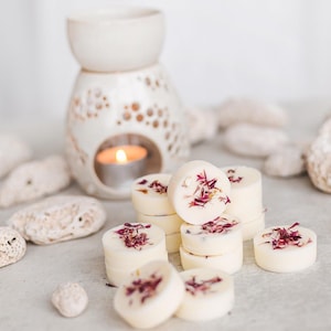 Puede incluir: Un difusor de aceite de cerámica blanca con una vela en el interior y una pila de fundidos de cera blanca con flores rosadas secas en la parte superior. Los fundidos de cera están dispuestos en forma de pirámide.