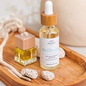May include: A wooden car diffuser with a string hanging from the top. The diffuser is filled with a clear liquid and has a wooden cap. The diffuser is sitting on a wooden tray with a small white rock next to it. The bottle is labelled "SHANTI naturals Wellbeing Car diffuser refill 100% natural with pure essential oils 30 ml shantinaturals.co.uk"