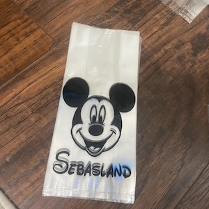 May include: Clear cellophane treat bags with a black and white Mickey Mouse graphic and the name "Sebasland" in black and blue lettering. The bags are folded and stacked on a wooden surface.