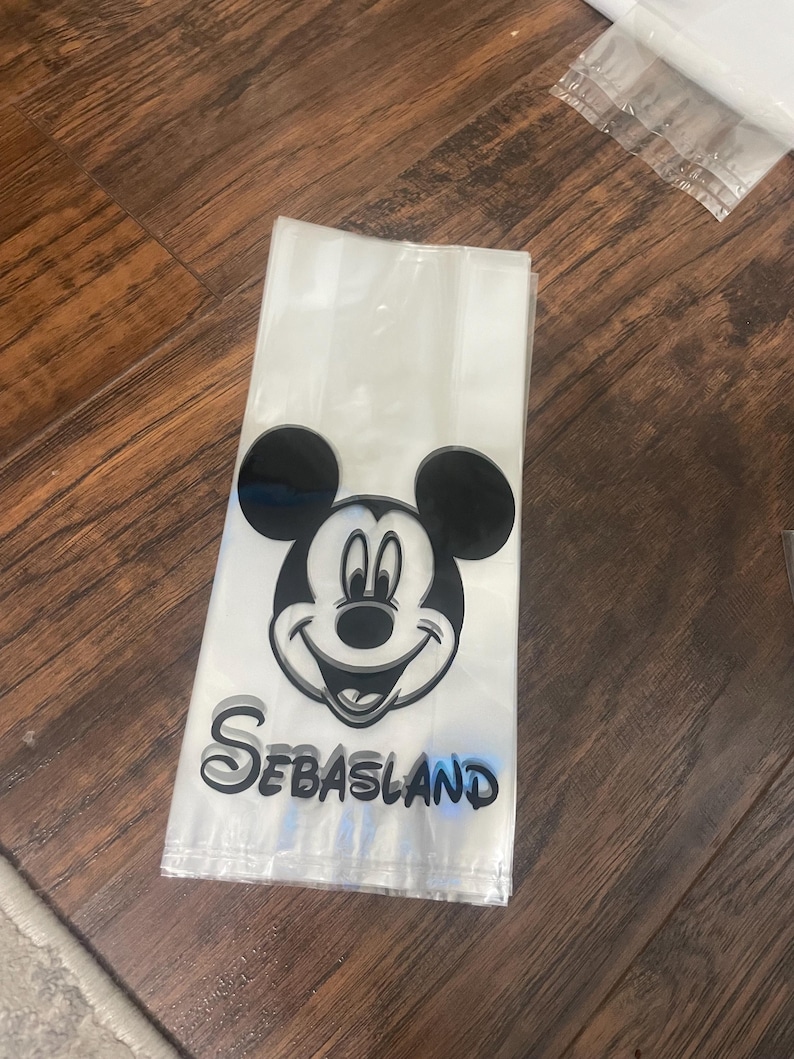 May include: Clear cellophane treat bags with a black and white Mickey Mouse graphic and the name "Sebasland" in black and blue lettering. The bags are folded and stacked on a wooden surface.