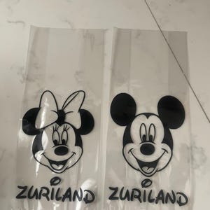 May include: Two clear plastic bags featuring black and white cartoon character faces. One bag has Minnie Mouse, the other has Mickey Mouse. The word "ZURILAND" is printed in black at the bottom of each bag.